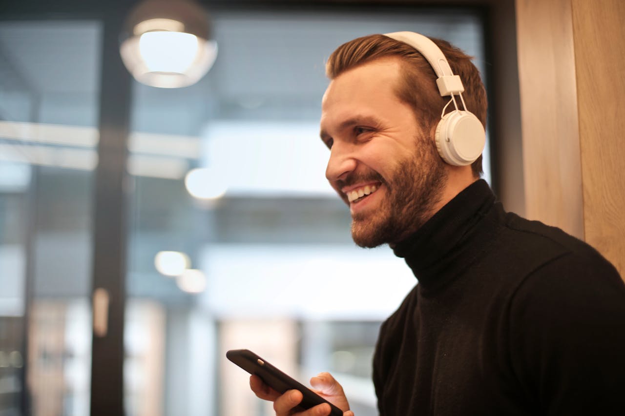 Bearded man smiling with headphones and smartphone indoors, enjoying music.