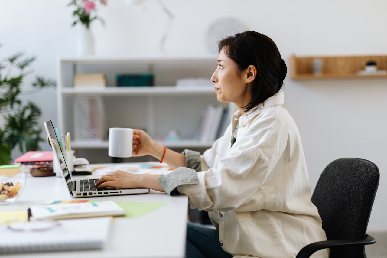 Asian woman working on laptop while holding a coffee cup in a modern office.